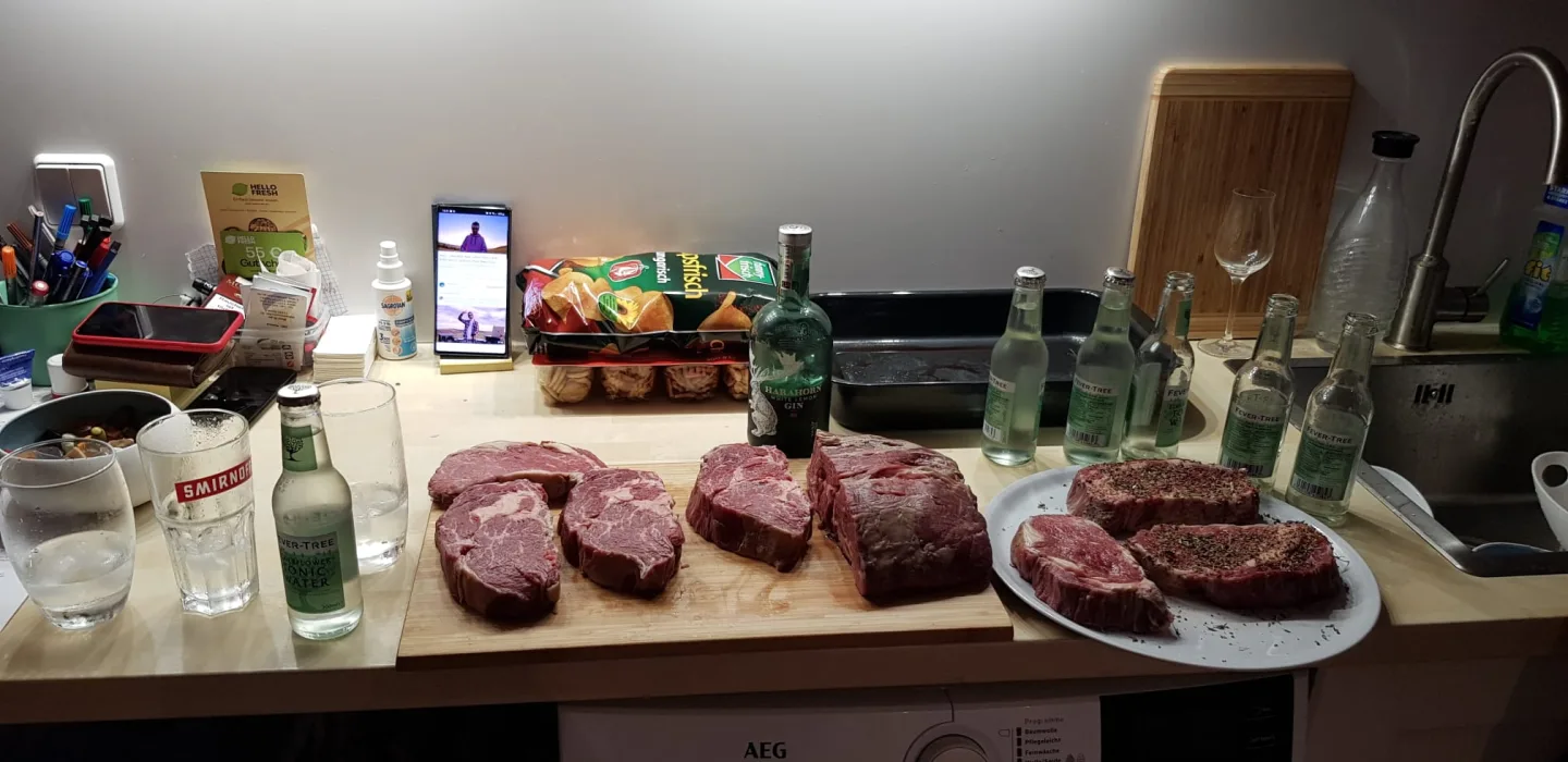 Fünf rohe Steaks auf Holzbrett mit Pfefferkruste, Gin und Tonic auf der Küchentheke