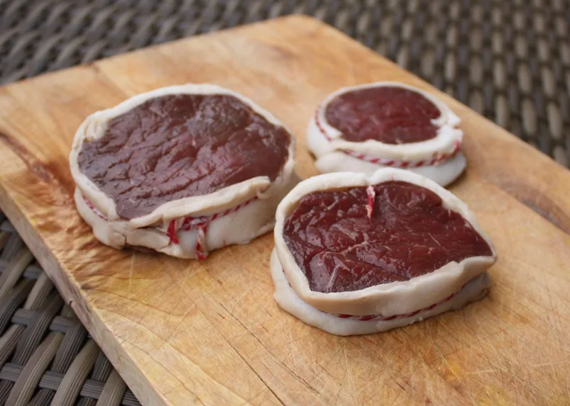 Drei rohe Hueftsteaks mit Speckmantel auf Holzbrett